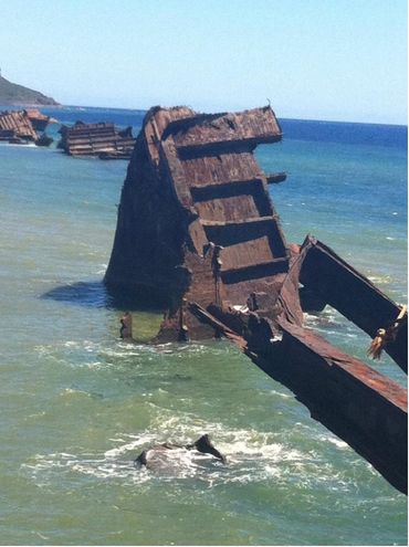 Rusted structural wreckage rising from shallow coastal water, with additional remains in the background.
